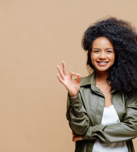 Indoor shot of pleasant looking curly woman has pleasant smile, makes okay gesture, excellent sign, gives approval, dressed in fashionable leather shirt, isolated over brown wall, blank space on left