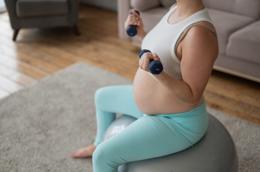 Pregnant woman doing exercises with dumbbells while sitting on a fitness ball at home. Close-up of a pregnant belly.