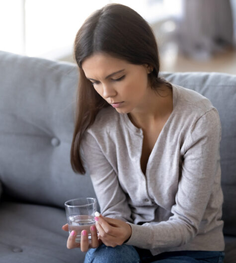 Alone woman sitting on sofa at home feels desperate thinking make decision termination of pregnancy looking at pill holding glass of water, depression pain relief, unwilling pregnancy abortion concept
