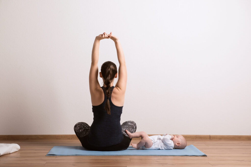 Woman sitting on a yoga mat with her back to the camera and her arms stretched above her head. Her baby is laying on the mat beside her.
