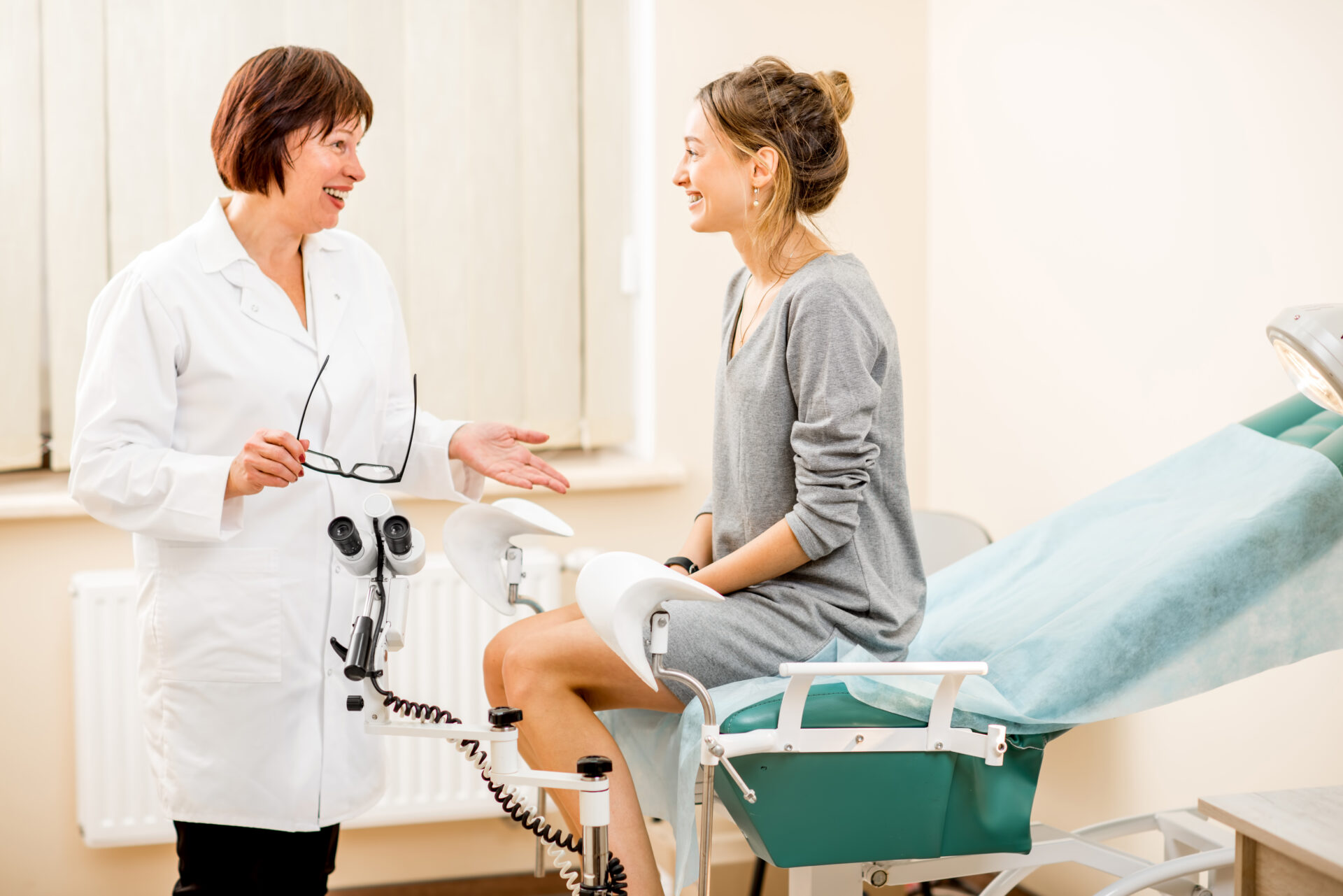 A woman is sitting on the exam table smiling as she speaks with her woman's healthcare provider.