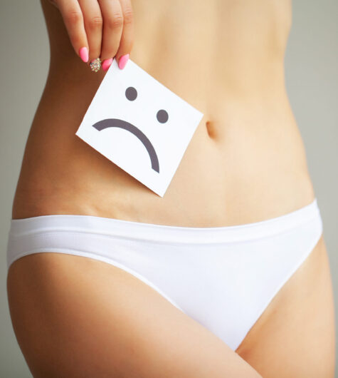 Closeup of a woman holding a piece of paper with a frowny face drawn on it. She is holding it near her pelvic area to signify poor vaginal health.
